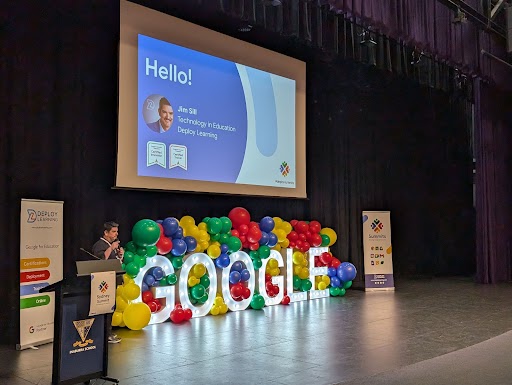 Jim Sill on stage with a screen and large lighted Google sign with balloons